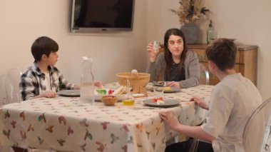 Samimi bir yemek salonunun geniş açılı görüntüsü ve kırsal-modern hissi. Bir anne ve iki çocuk öğle yemeğinden sonra masada otururlar. Çocuklar tabakları temizlemek için ayağa kalkar. Sıcak, günlük aile anları. Yatay, 4