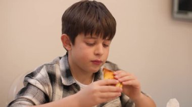 10 yaşındaki bir çocuğun, Latin Amerika tarzı bir börek yerken net bir zevkle çekilmiş fotoğrafı. Mutfak masasında oturmuş, memnun ve mutlu görünüyor. Ev yemeği, sıradan bir an. Yatay, 4K