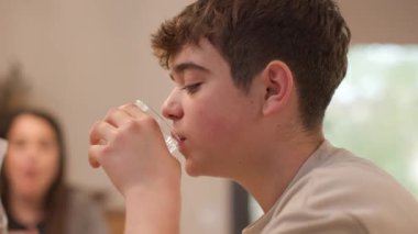 Açık kahverengi saçlı beyaz bir gencin aile yemeği sırasında hevesle bir bardak su içişini yakından izliyoruz. Annesi arka planda bulanık görünüyor. Yatay, 4K