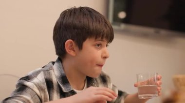 Öğle yemeği sırasında yemek masasında otururken bir bardak soğuk su içen koyu saçlı, beyaz bir çocuğun orta boy fotoğrafı. Arka planda bir televizyon görülebilir. Yatay, 4K