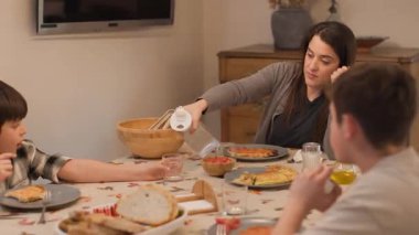 Evde yemek masasında oturan bir anne ve iki çocuğun orta boy fotoğrafları, yemeğe hazır. Onlar omlet, salata ve ekmekle sağlıklı bir Akdeniz yemeğinin tadını çıkarmaya hazırlanırken, o su koyuyor. Yatay, 4K