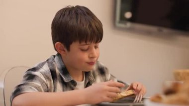Koyu saçlı beyaz bir çocuğun evde öğle yemeği sırasında ailesiyle sohbet ederken bir dilim domatesli ekmek yerken orta boy fotoğrafı. Arka planda televizyon olan sıcak bir aile ortamı. Yatay, 4K