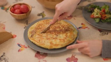 Bir İspanyol omletini yemek masasında porsiyonlar halinde keserken çekilmiş yüksek açılı bir fotoğraf. Masada görünüyor: ekmek için salata ve olgun domates. Otantik, günlük yemek sahnesi. Yatay, 4K
