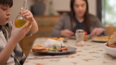 Evde yemek masasında ailesiyle yemek yerken salatasına zeytinyağı ve tuz ekleyen bir çocuğun orta boy fotoğrafı. Anne arka planda bulanık görünüyor. Günlük sağlıklı rutin. Yatay 4K