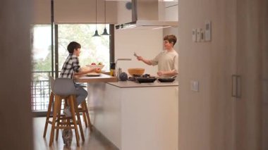 Parlak, kırsal ve modern bir mutfaktaki bir ailenin geniş açılı görüntüsü. Çocuklar sofrayı hazırlamak için bulaşıkları götürerek yardım ederler. Geniş bir evde sıcak, günlük aile ilişkileri. Yatay, 4K
