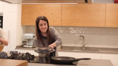 Geniş bir mutfakta bir anne tavadan bir İspanyol omletini tabağa fırlatıyor, sonuçtan açıkça memnun görünüyor. Oğlu yanında kutlama yapıyor. Parlak, gerçek bir aile yemeği anı. Yatay, 4K