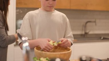 Mutfakta salata hazırlayan 14 yaşındaki bir çocuğun orta boy fotoğrafı. Annesine aile yemeğinde yardım ediyor. Gerçek bir ev içi sorumluluk anı. Yatay, 4K