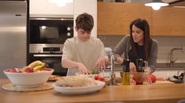 Bir anne ve iki çocuğunun mutfak adasında salata ve sağlıklı yiyecekler hazırlarken geniş cepheli görüntüleri. Doğal ışık, kırsal-modern alanı doldurur. Gerçek bir aile işbirliği. Yatay, 4K