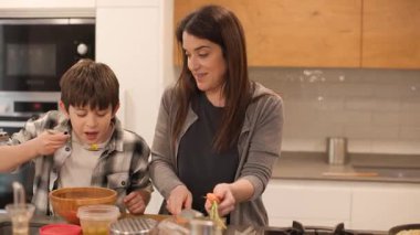 Mutfakta sıcak, eğlenceli bir sahne: 10 yaşındaki bir çocuk, annesi salata için sebze doğrarken kaseden zeytin alıyor. Sevgi ve bağlılık anında birlikte gülerler. Yatay, 4K
