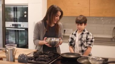 10 yaşındaki oğluna İspanyol omleti yapmayı öğreten bir annenin geniş açılı görüntüsü. Patates ve yumurta karışımını tavaya döküyorlar. Geniş, modern bir mutfakta doğal ışık. Yatay, 4K