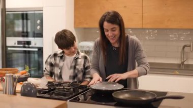 10 yaşındaki oğluna İspanyol omletinin nasıl tabağa konulacağını gösteren bir anne resmi. Doğal ışıkla dolu, büyük, modern bir mutfakta aile anı paylaştık. Yatay, 4K