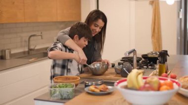 Bir anne ve oğlunun mutfak adasında yemek pişirme anlarını geniş açılardan görürler. Çocuk ev işlerini öğrenirken metal bir kasede yumurta çırpıyor. Parlak modern mutfak. Yatay, 4K