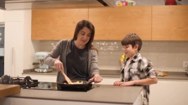 Bir anne ve 10 yaşındaki oğlunun mutfak adasındaki bir tavada yemek pişirirken konuştuğu orta boy bir fotoğraf. Günlük anlarımızı ve aşçılık bilgimizi ev mutfağında paylaşıyoruz. Yatay, 4K