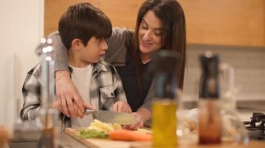 Bir annenin, patatesleri bıçakla keserken 10 yaşındaki oğluna rehberlik ettiği orta büyüklükte bir fotoğraf. Güvenliği sağlamak için yakından denetliyor. Gerçek bir aile öğrenme ve bakım anı. Yatay, 4K