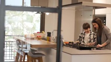 Geniş açı mutfak kapısından görülüyor: annesi sebzeleri nasıl keseceğini açıklarken gülümseyen bir çocuğun yakından dinlediği modern, güneşli bir mutfak. Sessiz bir öğrenme ve kaynaşma anı.