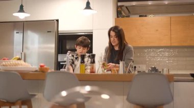 Modern bir mutfak adasında bir anne ve oğlunun omuz omuza yemek pişirdiği geniş bir cephe fotoğrafı. Çocuk sebze yıkayarak, ev işlerini doğal ışık altında paylaşarak yardımcı oluyor. Yatay, 4K