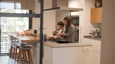 Mutfak kapısından geniş açı görünüyor: geniş, modern, güneşli bir mutfak ve adada bir anne ve oğlu var. Çocuk salata için havuç yıkamaya yardım ediyor. Tabureli bir bar görülebilir. Yatay, 4K