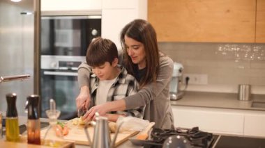 Bir annenin 10 yaşındaki oğluna mutfak adasında bıçakla nasıl güvenli bir şekilde patates kesileceğini öğrettiği geniş açı. Modern ev mutfağı ve ortak aile öğrenme anı. Yatay, 4K