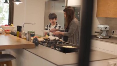 Mutfak kapısından geniş açı görünüyor: anne ve oğlunun adada birlikte yemek pişirdiği parlak, modern bir mutfak. O, ona sebzeleri güvenli bir şekilde nasıl keseceğini öğretirken gülümsüyor. Yatay