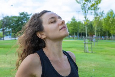 Young woman relaxing, feeling alive, breathing fresh air, calm and dreaming with closed eyes, in public park