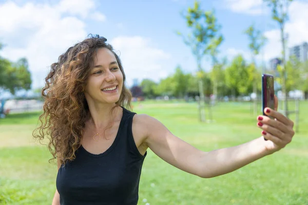 Young woman with curly brunette hair doing selfie on summer day in park