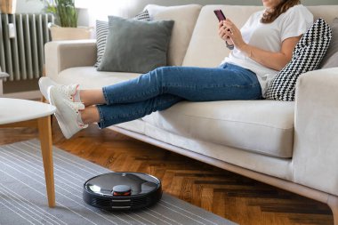 Robot elektrikli süpürge oturma odasını temizliyor. Genç bayan dinleniyor, evdeki kanepede oturuyor.