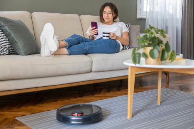 Robot elektrikli süpürge oturma odasını temizliyor. Genç bayan dinleniyor, evdeki kanepede oturuyor.