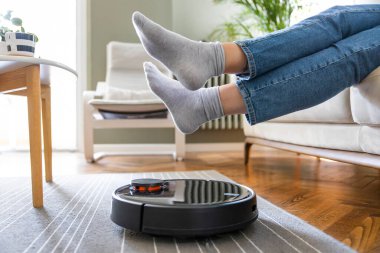 Genç kadın robot süpürgenin geçmesine izin verdiği için ayaklarını kaldırıyor.
