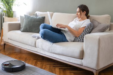 Robot elektrikli süpürge oturma odasını temizliyor. Genç bayan dinleniyor, evdeki kanepede oturuyor.