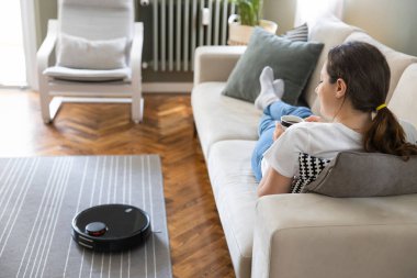Robot elektrikli süpürge oturma odasını temizliyor. Genç bayan dinleniyor, evdeki kanepede oturuyor.