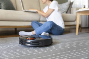 Robot vacuum cleaner cleaning the living room. Young woman enjoy rest, sitting on sofa at home