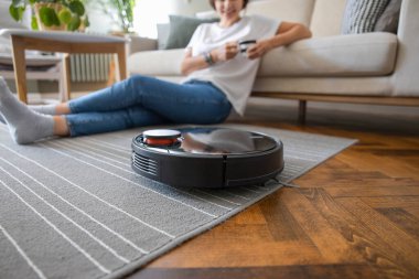 Robot elektrikli süpürge oturma odasını temizliyor. Genç bayan dinleniyor, evdeki kanepede oturuyor.