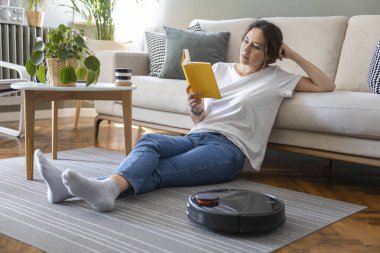 Robot elektrikli süpürge oturma odasını temizliyor. Genç bayan dinleniyor, evdeki kanepede oturuyor.