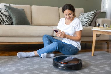 Robot vacuum cleaner cleaning the living room. Young woman enjoy rest, sitting on sofa at home