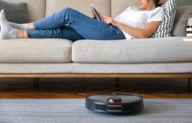 Robot vacuum cleaner cleaning the living room. Young woman enjoy rest, sitting on sofa at home