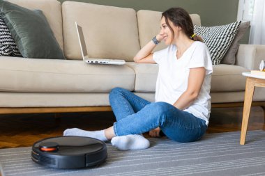 Robot elektrikli süpürge oturma odasını temizliyor. Genç bayan dinleniyor, evdeki kanepede oturuyor.