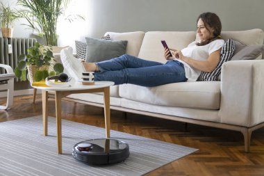 Robot elektrikli süpürge oturma odasını temizliyor. Genç bayan dinleniyor, evdeki kanepede oturuyor.