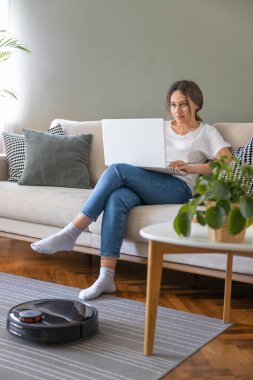 Robot vacuum cleaner cleaning the living room. Young woman enjoy rest, sitting on sofa at home