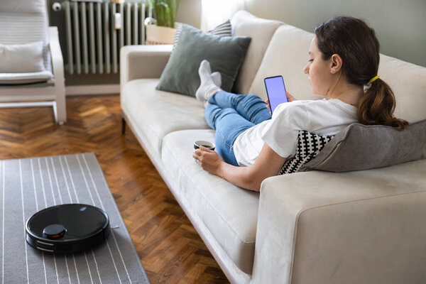 Робот-пылесос убирает гостиную. Молодая женщина наслаждается отдыхом, сидя на диване дома