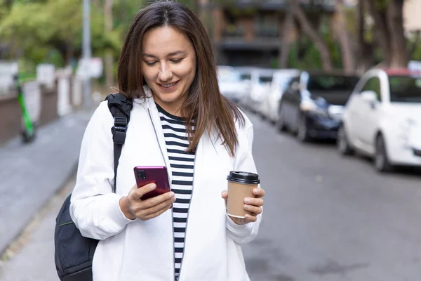 여자가 스마트폰으로 행복하게 마시면서 시내에서 커피를 빼앗아 — 스톡 사진