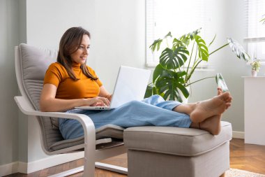 Evde dizüstü bilgisayar kullanan genç bir kadın. Ev işi veya çevrimiçi alışveriş, uzaklık öğrenme, eğitim çevrimiçi, e-ticaret, serbest danışma