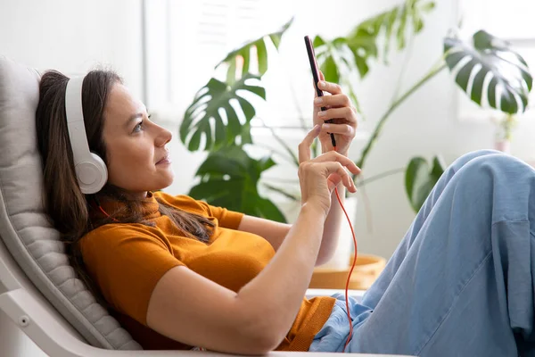 自宅の携帯電話からヘッドフォンで音楽を聴く魅力的な若い女性 — ストック写真