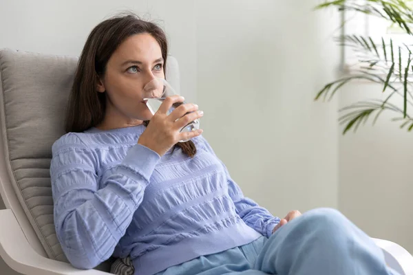 年轻妇女坐在家里的椅子上喝水 饮水充足概念对健康的好处 — 图库照片