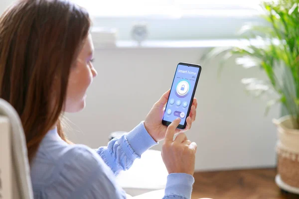 Young woman holding phone with app smart home on the screen in the room. Concept of controlling home security from a mobile devic