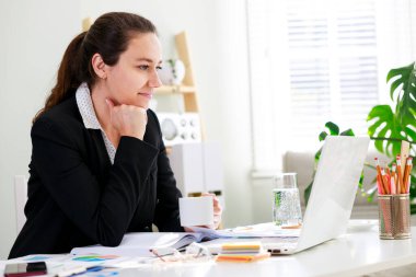 Evde dizüstü bilgisayarla çalışırken kahvesinin tadını çıkaran genç bir kadın.