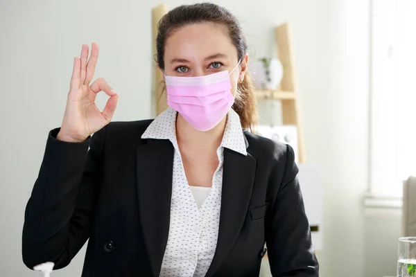 Business woman with face mask gesturing ok sign avoiding coronavirus at home office