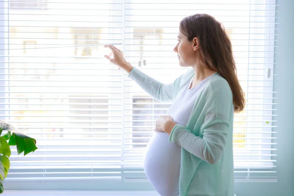 幸せな妊娠中の若いです女性タッチ彼女の妊娠中の腹と立ちますザ ウィンドウ — ストック写真