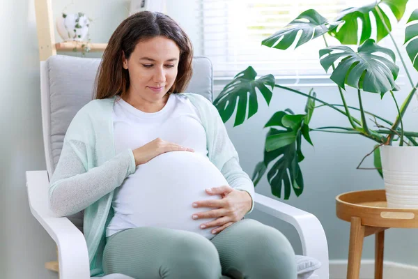 幸せな妊娠中の若いです女性タッチ彼女の妊娠中の腹と座っている窓の近く — ストック写真