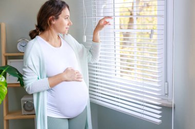 Mutlu hamile kadın hamile karnına dokunuyor ve pencerenin kenarında duruyor. 