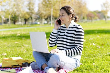 Parkta dizüstü bilgisayarıyla oturan ve telefonla konuşan genç bir kadın.